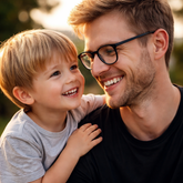 Padre joven rubio con gafas negras y camiseta negra sonríe mientras abraza a su hijo pequeño de 4 años, también rubio, en un momento de complicidad y felicidad al aire libre con luz cálida de atardecer